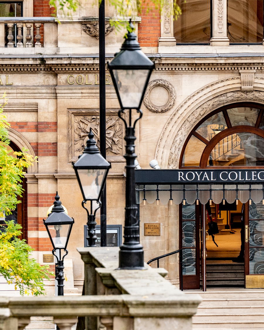 The entrance of the Royal College of Music, framed by black lampposts and greenery on the left, with an open door.